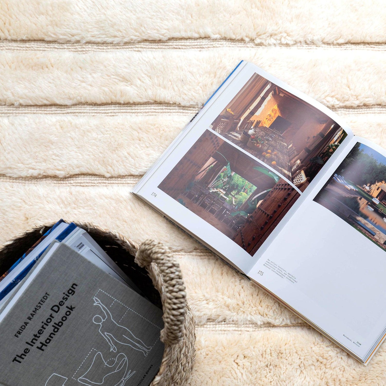 An open book with vivid interior design photos placed on a soft, cream-colored wool rug from Barbary Wool, alongside a woven basket filled with design handbooks.