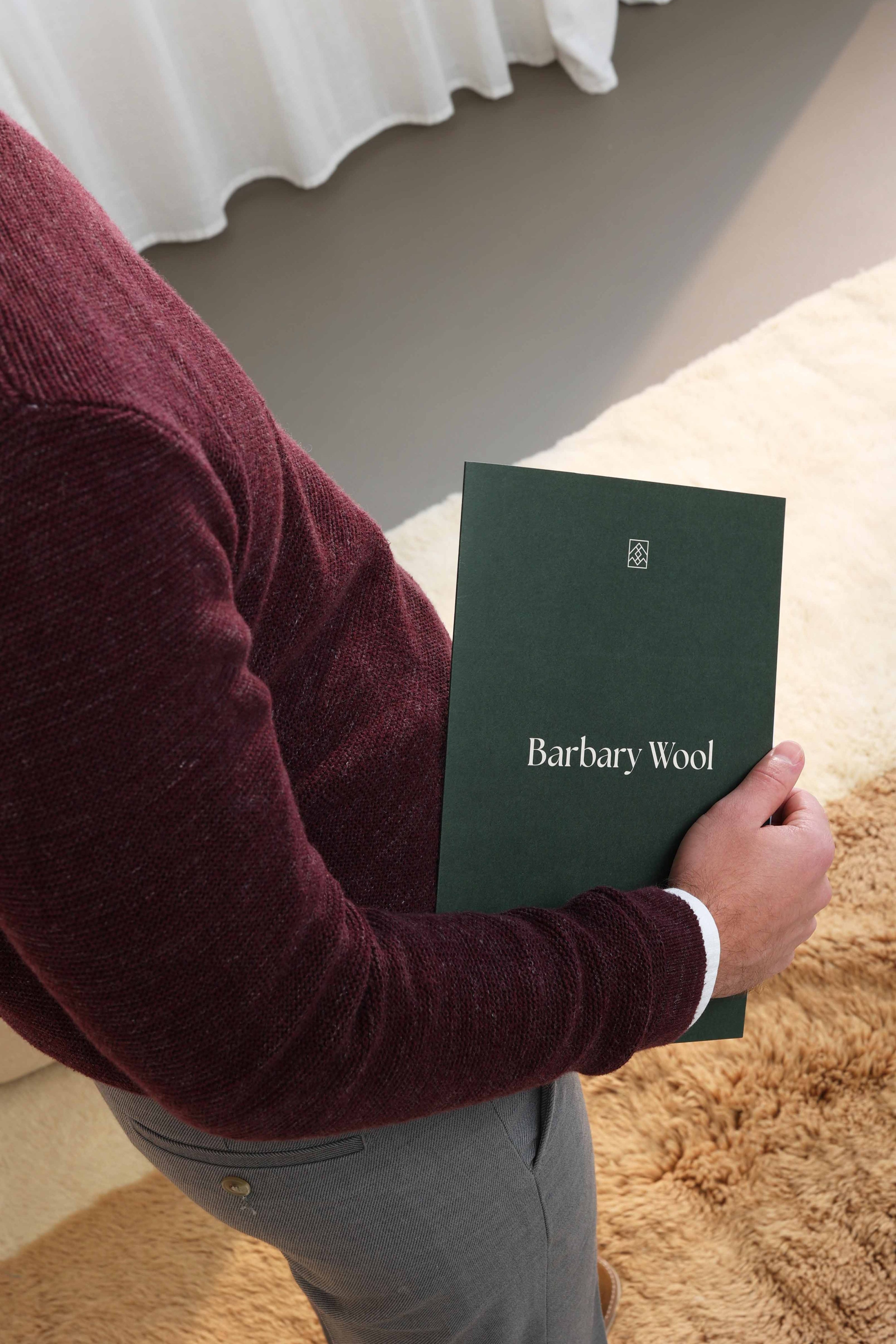 Man holding a dark green Barbary Wool catalog while standing on a soft beige rug in a cozy indoor setting, wearing a maroon sweater and gray trousers.