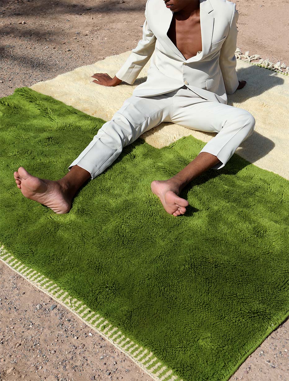 A person in a light beige suit sitting on a Barbary Wool rug with a green and cream two-tone design, placed outdoors on a sandy surface, highlighting the rug's vibrant color and soft texture.