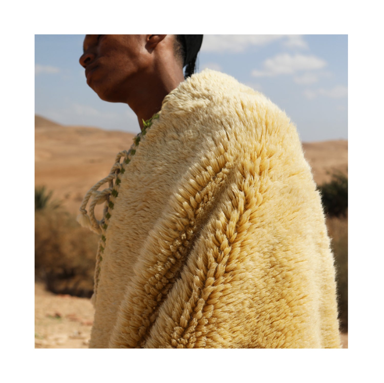 A person draped in a luxurious, handwoven wool rug with a golden-yellow hue, standing in a desert landscape under a bright blue sky, evoking warmth and natural craftsmanship.