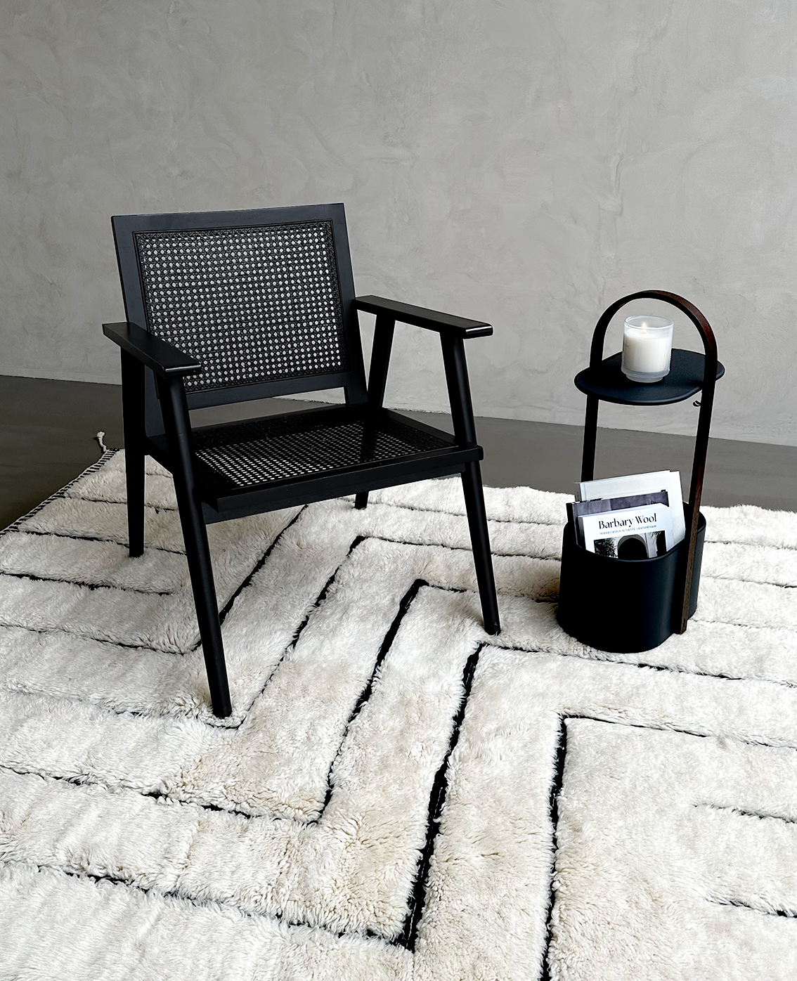 A cozy setup featuring a black rattan chair and a modern side table holding a candle and a Barbary Wool magazine, all placed on a luxurious white wool rug with black geometric patterns.