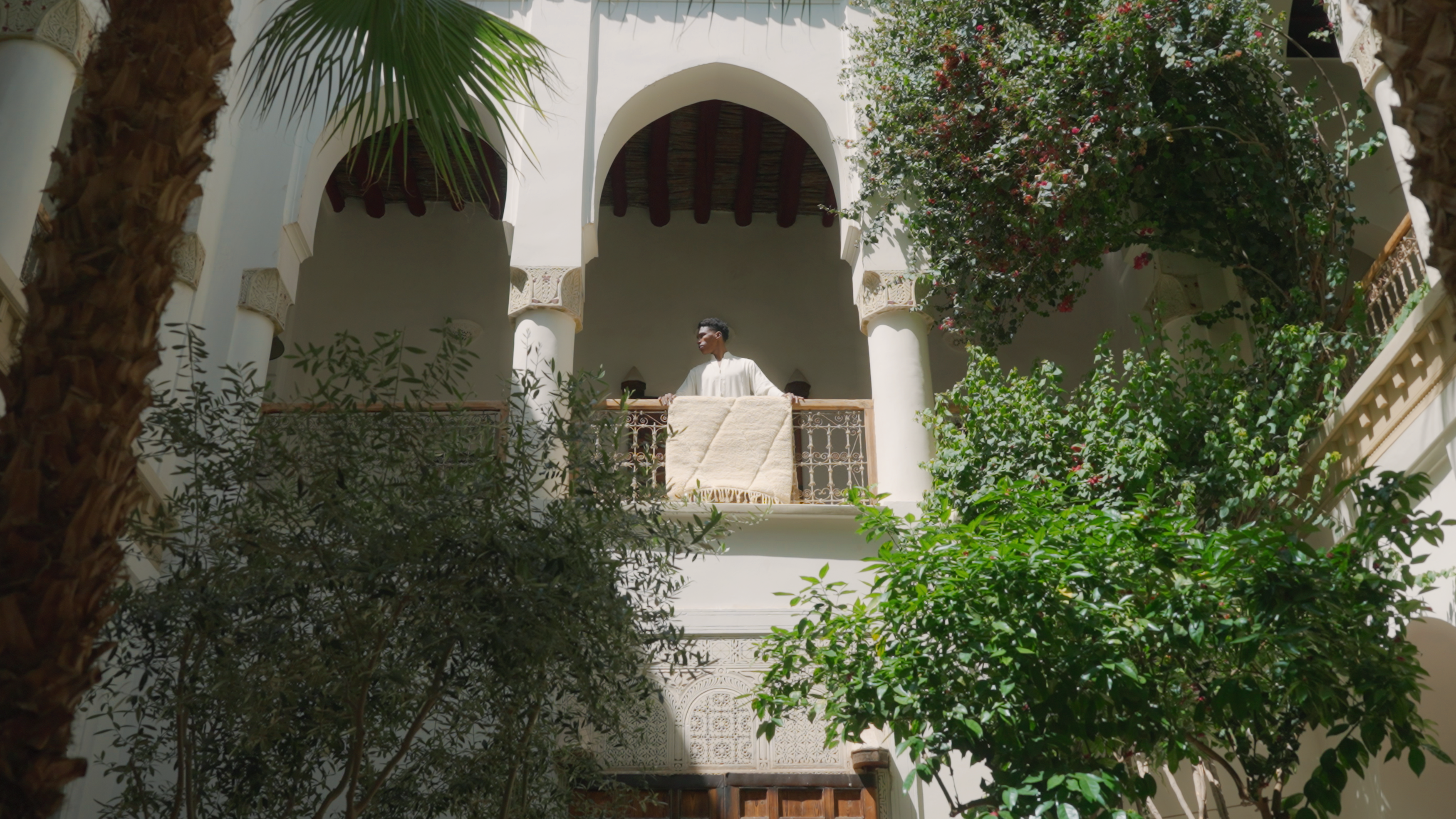 Person standing on a balcony of a Moroccan-style building, holding a wool rug made with natural materials, surrounded by lush greenery, promoting Barbary Wool handcrafted rugs.