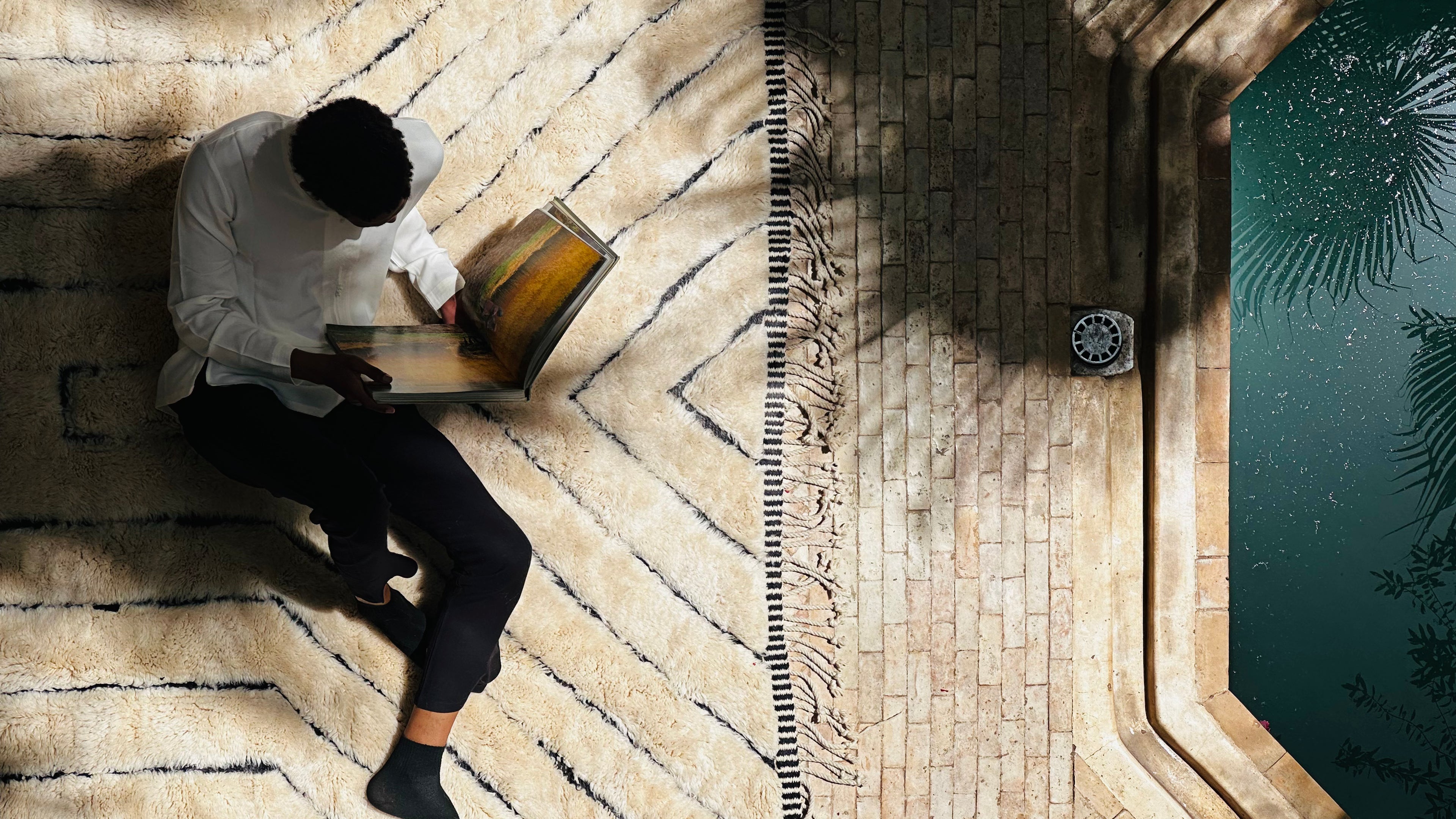 Person sitting on a luxurious wool rug with geometric patterns, reading a book in natural light, showcasing the comfort and style of Barbary Wool's handcrafted rugs.