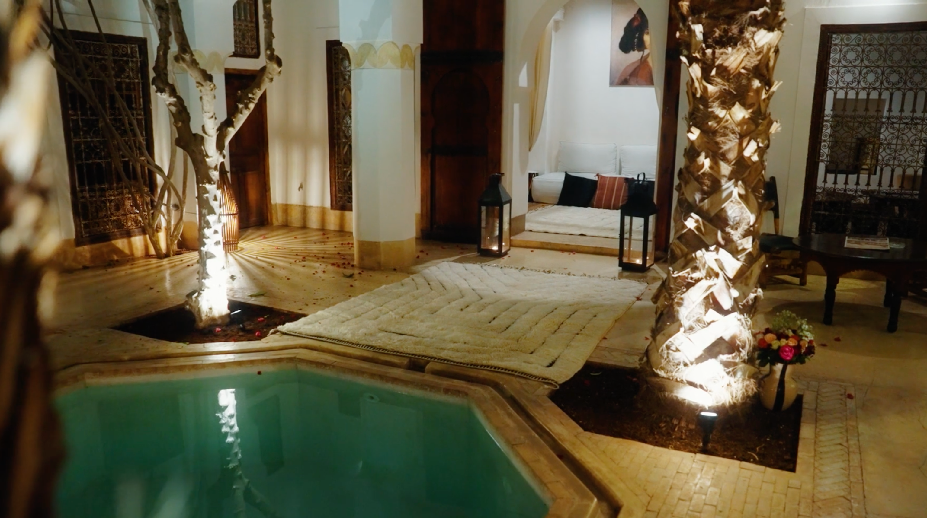 A serene courtyard setting featuring a softly lit pool surrounded by elegant architecture. The space includes a textured cream rug from Barbary Wool, accenting the natural tones of the decor. Illuminated palm trees, intricate metalwork, and a cozy seating area with cushions and lanterns create a warm and inviting ambiance.