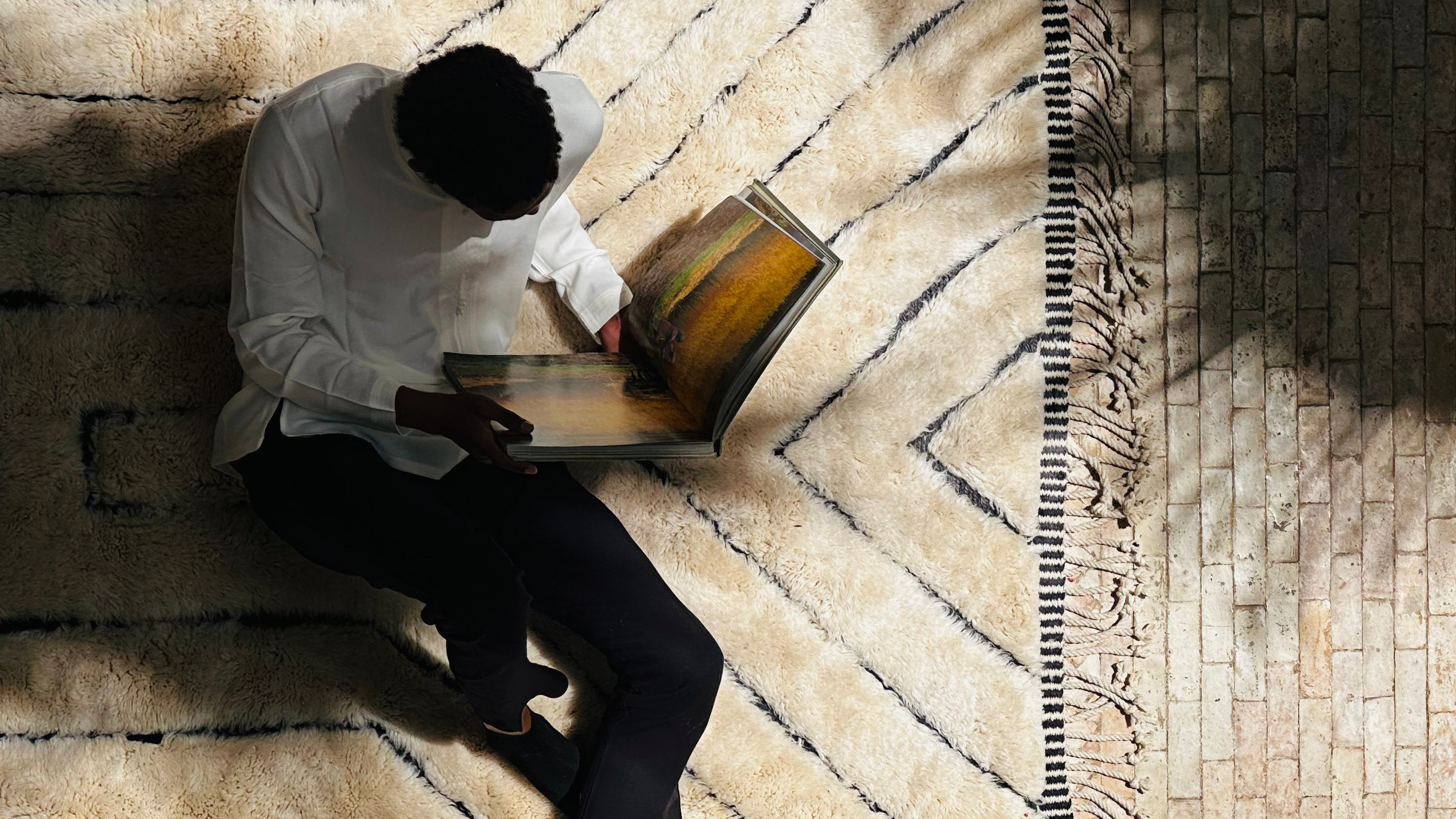 An individual wearing a white shirt and dark pants sits on a textured cream and black-patterned rug while reading a book. The scene is warmly lit, with soft sunlight casting shadows across the rug and nearby brick flooring, creating a cozy and serene atmosphere.