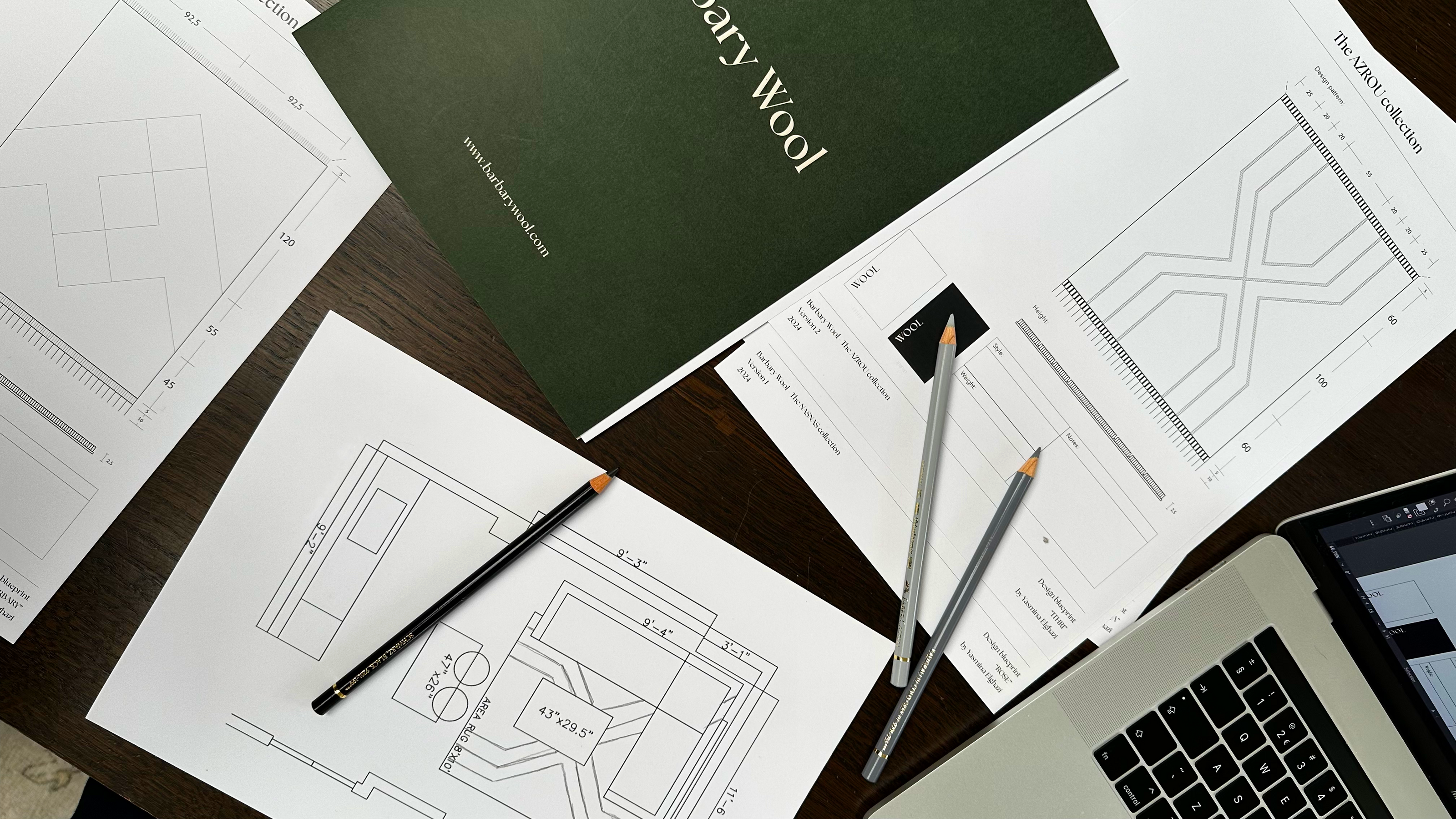 A workspace showcasing design materials from Barbary Wool's, including detailed floor plans, diagrams, and a green booklet labeled 'Barbary Wool.' The setup features pencils, architectural drawings, and a laptop, emphasizing a focus on creative planning and design.